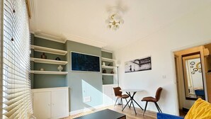 A living room with a blue sofa, a black coffee table, a dining table with chairs, and a white cabinet with shelves. - Stylish Kennington flat w/garden, KING bed nr tube (Greater London)
