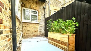A narrow courtyard with a wooden planter box filled with plants, a brick wall, and a black fence. - Stylish Kennington flat w/garden, KING bed nr tube (Greater London)