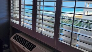 Ein Zimmer mit großem Fenster, horizontalen Jalousien, einer Wand-Klimaanlage und Blick auf die Küste.