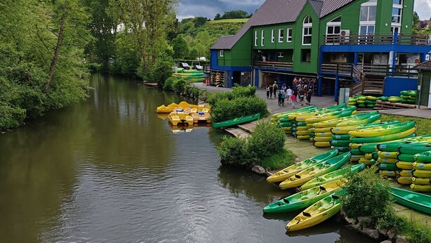A riverside area with green and yellow canoes lined up near buildings and a walkway. - Au Sein de la Suisse Normande, Randonnée, Vélo et le Kayak Pour Tous (Pont-d'Ouilly)