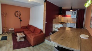 A compact living space with a sofa, a wooden dining table, a kitchen area with cabinets and a stove, and a wall clock. - Au Sein de la Suisse Normande, Randonnée, Vélo et le Kayak Pour Tous (Pont-d'Ouilly)