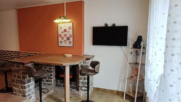 A cozy living area with a brick fireplace, a wooden dining table, and two bar stools. A flat-screen TV is mounted on the wall, and there's a small shelf with decorative items.