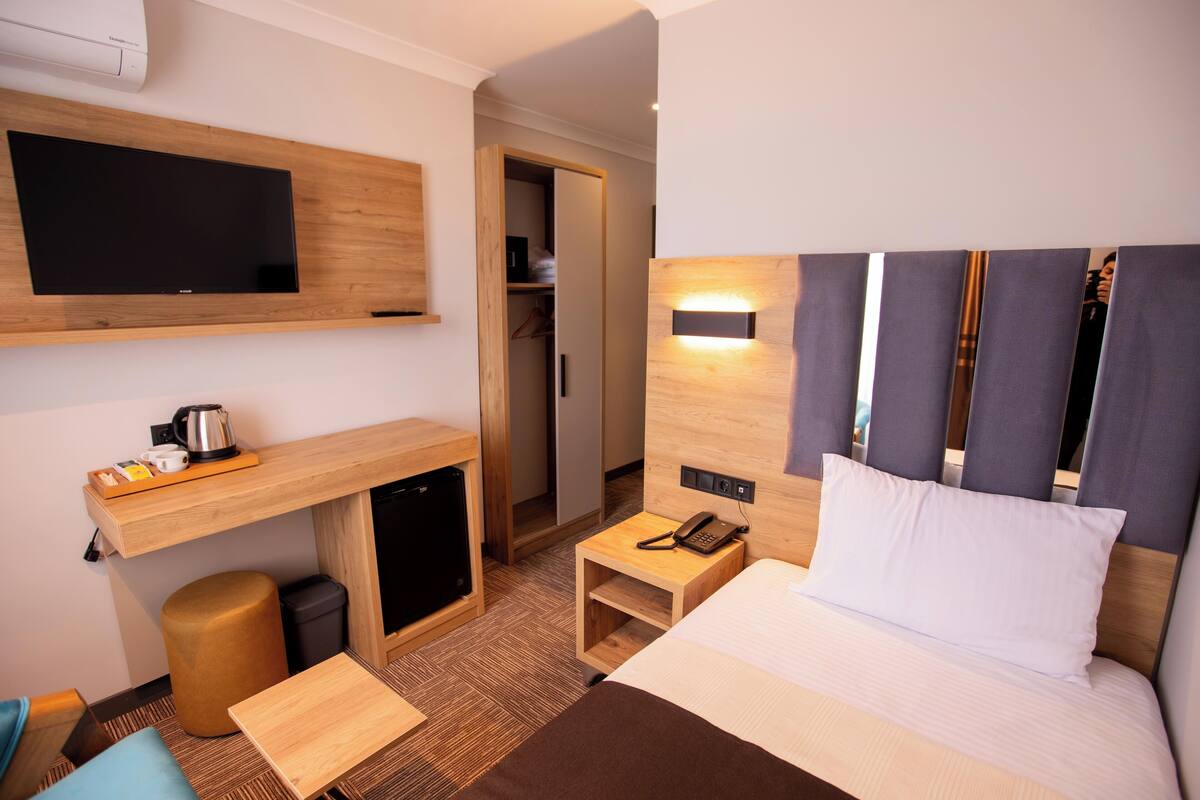 A hotel room with a bed, a desk, a chair, a small table, a trash bin, a telephone, and a television mounted on the wall.