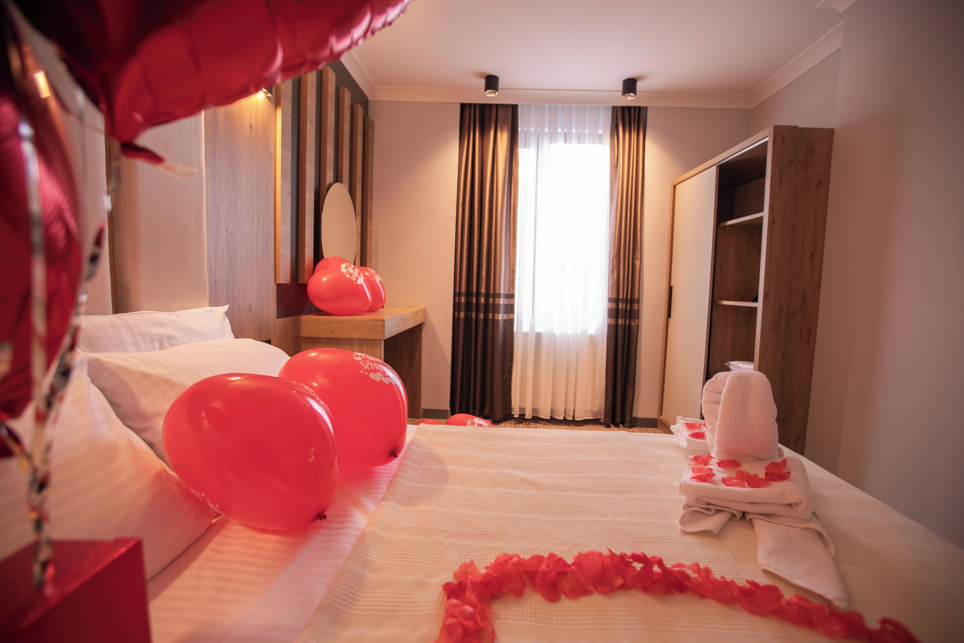 A hotel room with a neatly made bed, a bedside table with a red balloon, a mirror, and a wardrobe.