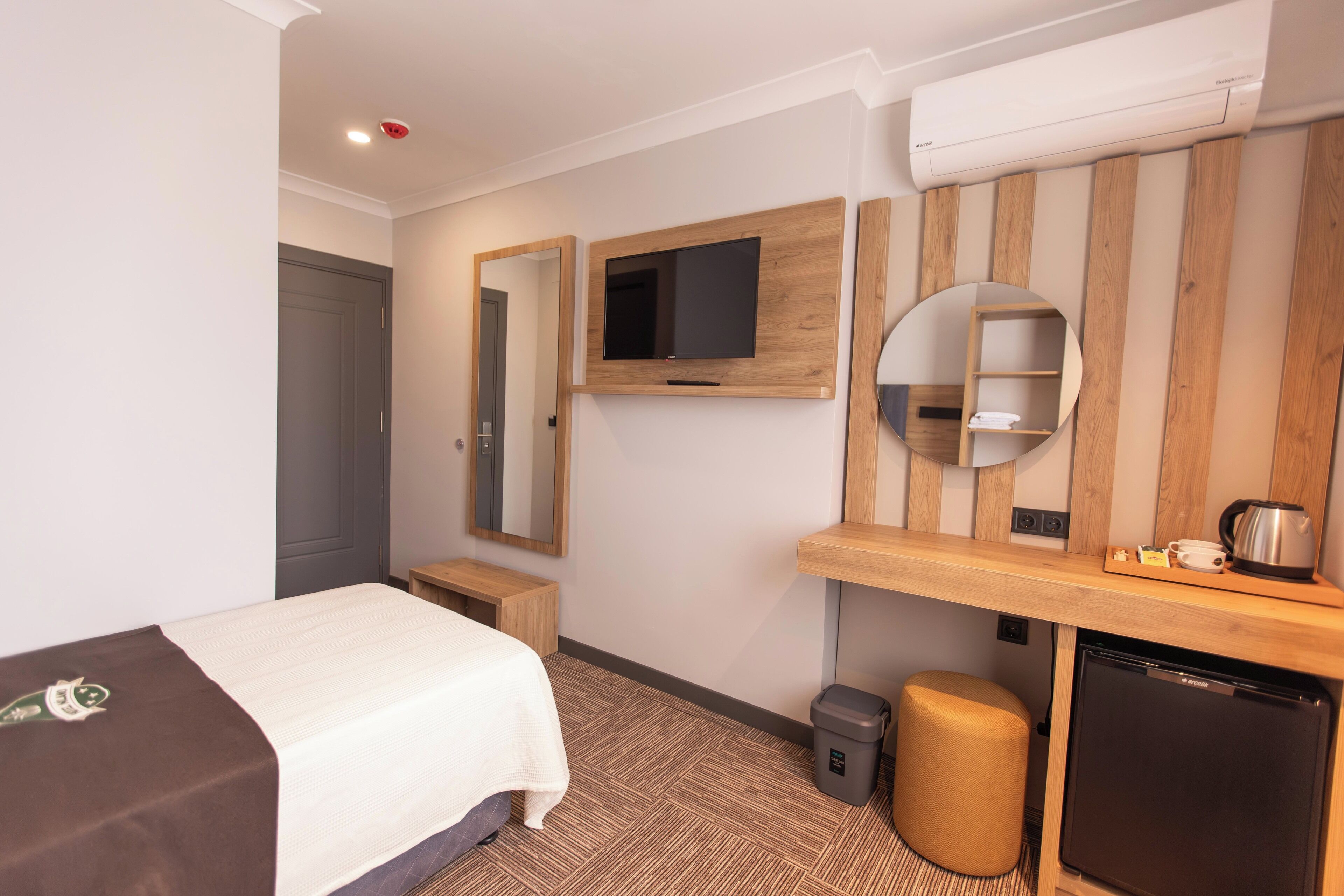 Habitación de hotel con cama, televisor montado en la pared, un pequeño escritorio con hervidor y un cubo de basura.