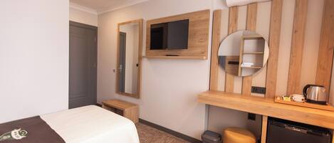 Habitación de hotel con cama, televisor montado en la pared, un pequeño escritorio con hervidor y un cubo de basura.