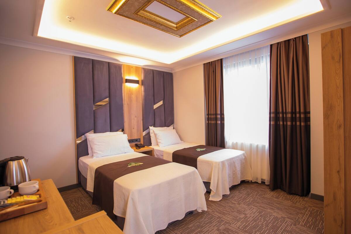 A hotel room with two single beds, a wooden headboard, a bedside table with a kettle, and a large window with curtains.