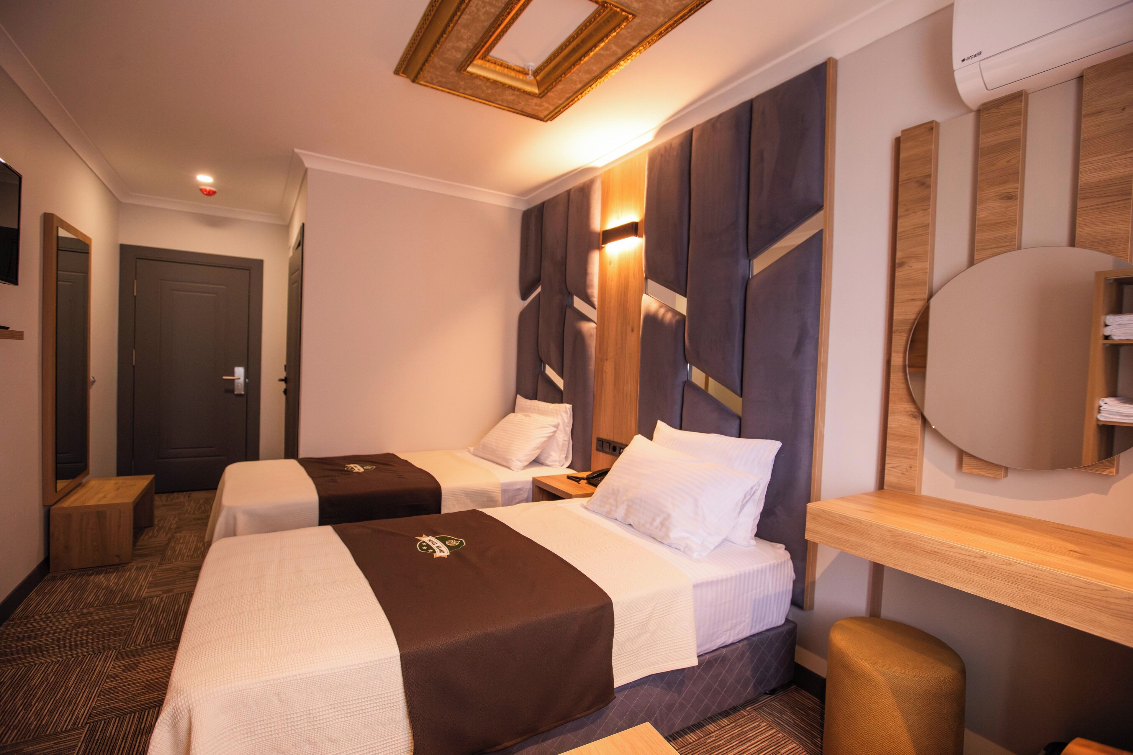 A hotel room with two beds, a wooden headboard, a bedside table, a small stool, and a large mirror.