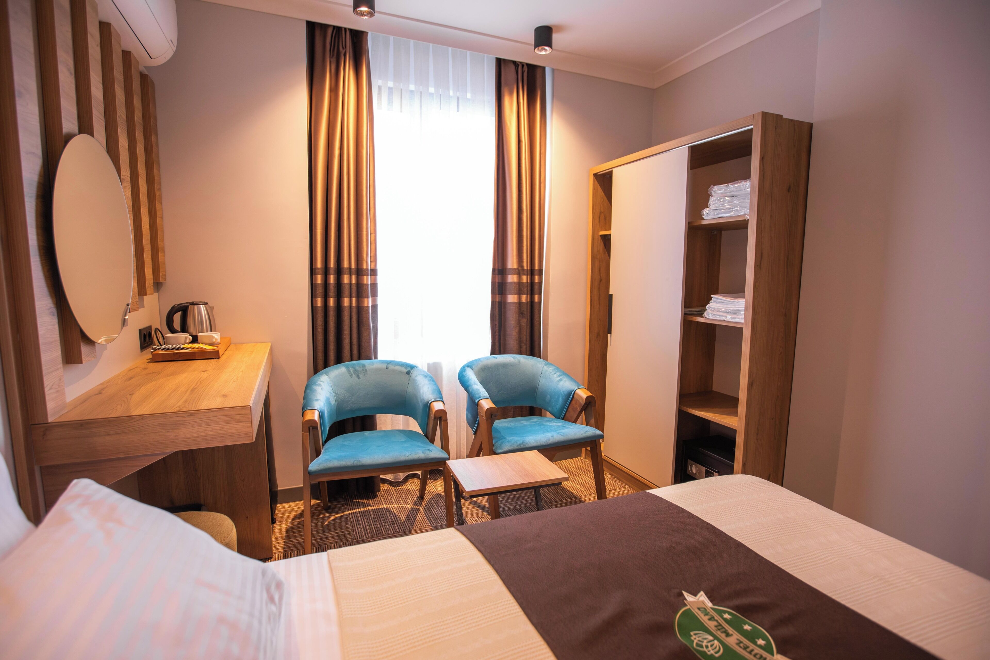 A hotel room with a bed, two blue chairs, a wooden desk with a coffee pot, a mirror, and a wardrobe.