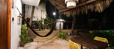 A patio area with a wooden table and yellow chairs, a hanging hammock, and a thatched roof structure.