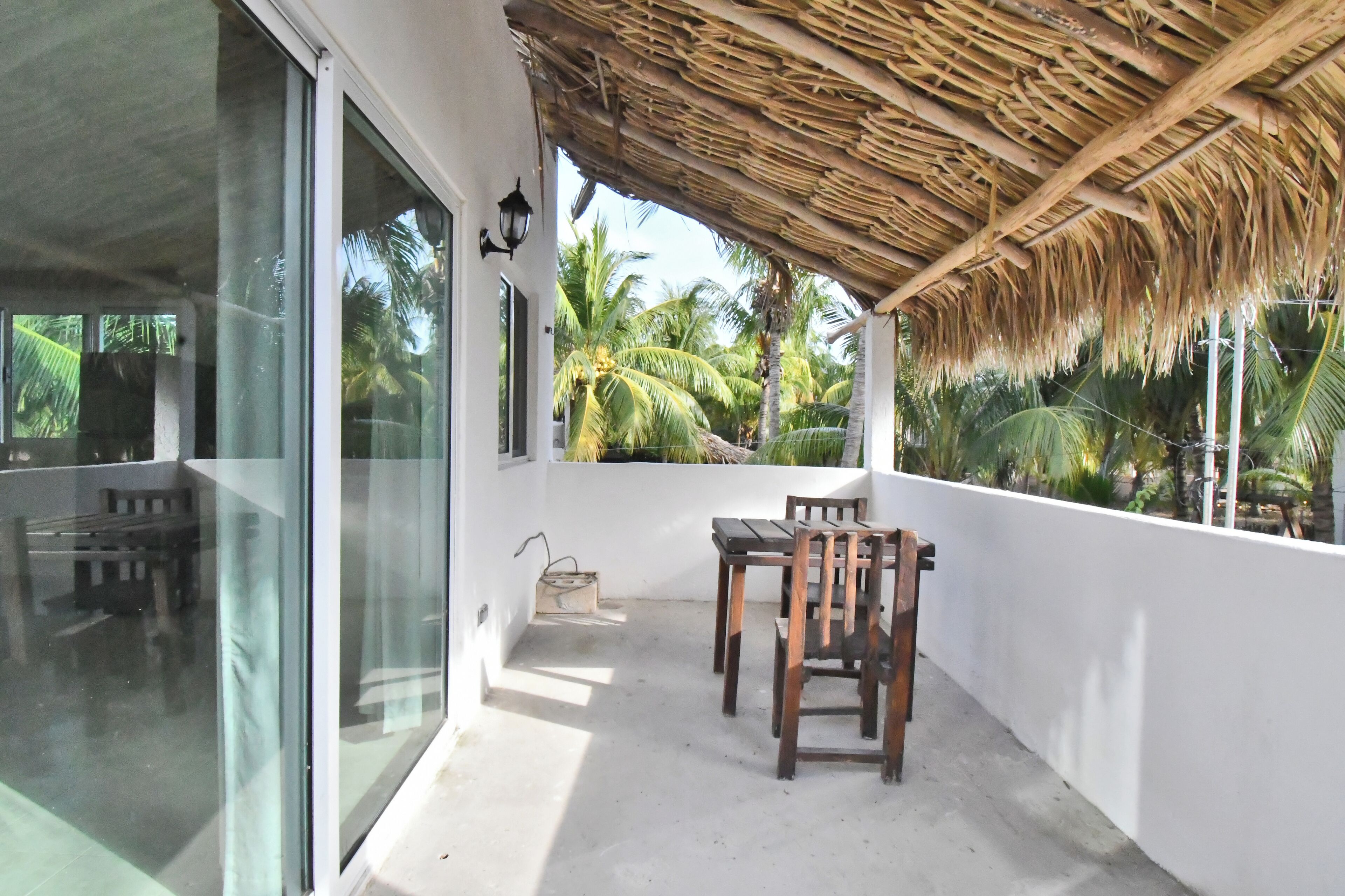 A balcony with a thatched roof, a table with chairs, and a view of palm trees.