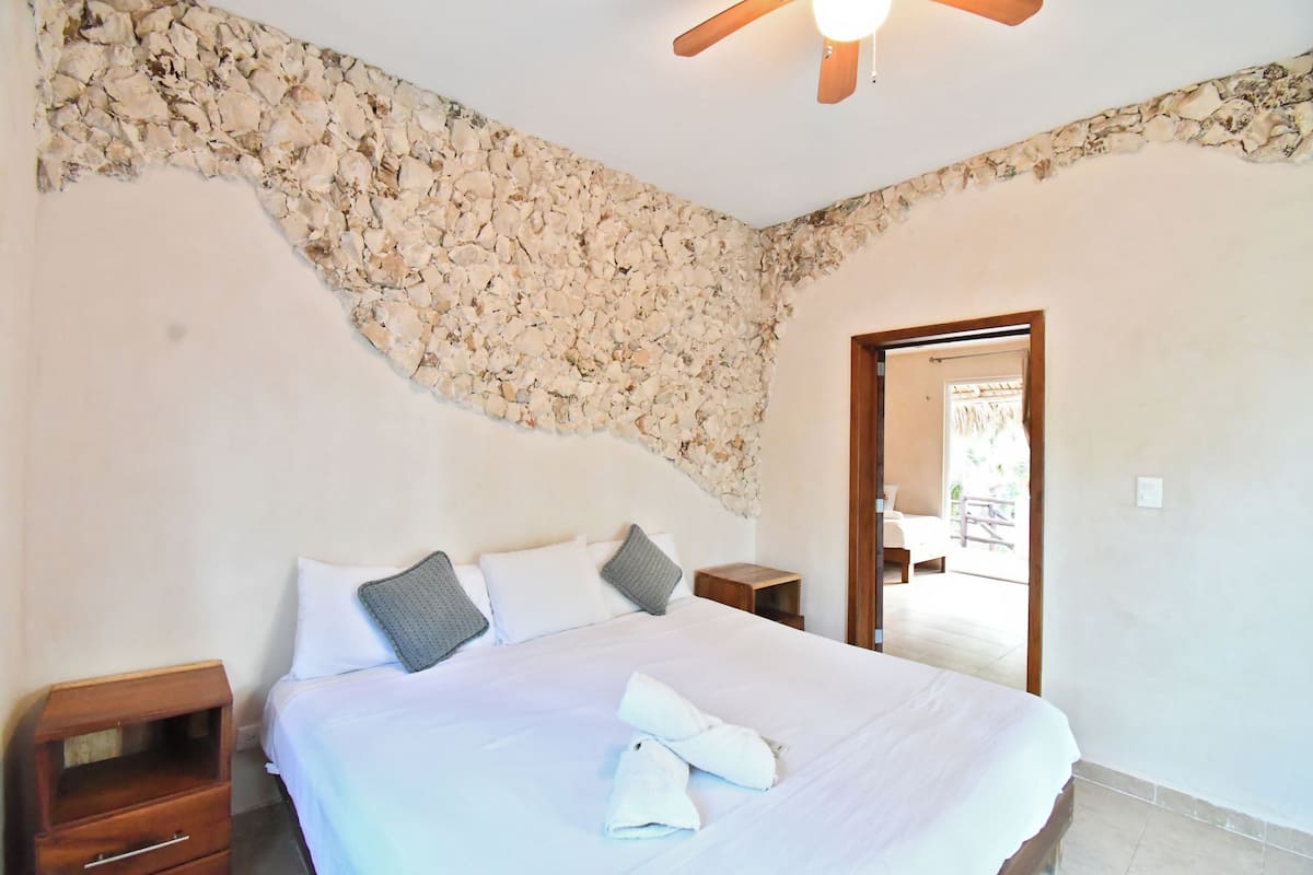 Un dormitorio con una pared de acento de piedra, una cama con ropa de cama blanca, dos almohadas, una mesita de noche de madera y un ventilador de techo.