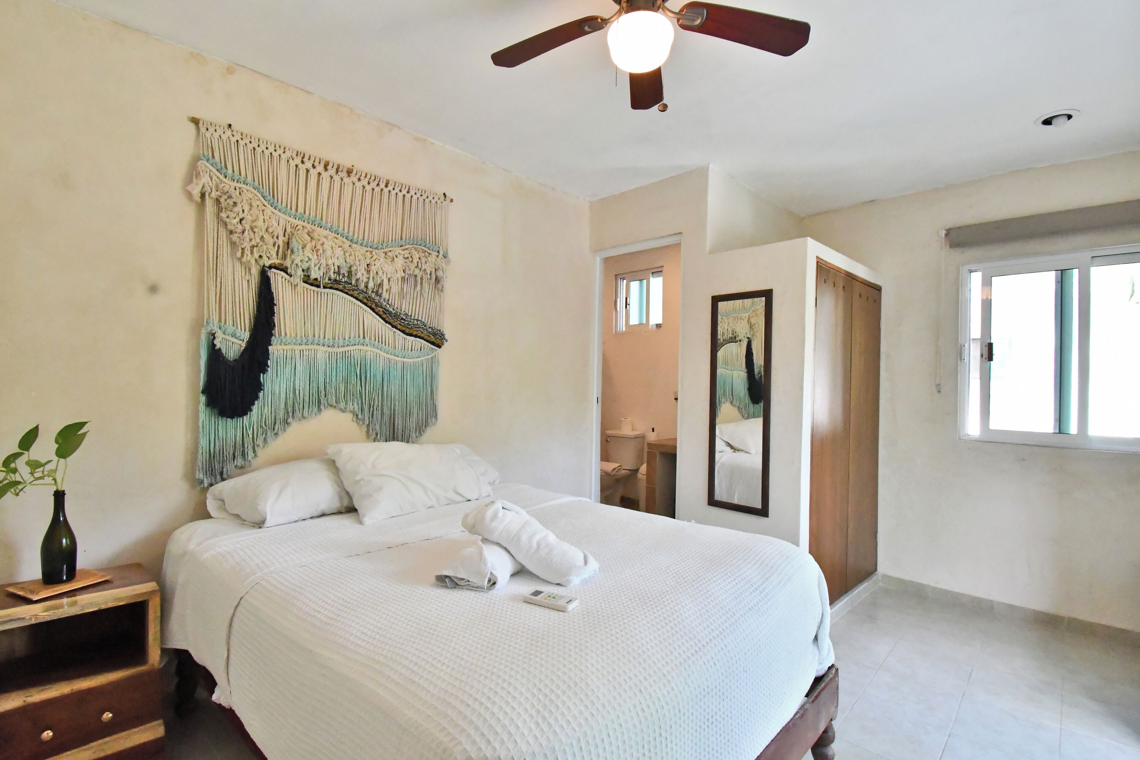 A bedroom with a bed, a ceiling fan, a wall tapestry, a bedside table with a vase, and a window.