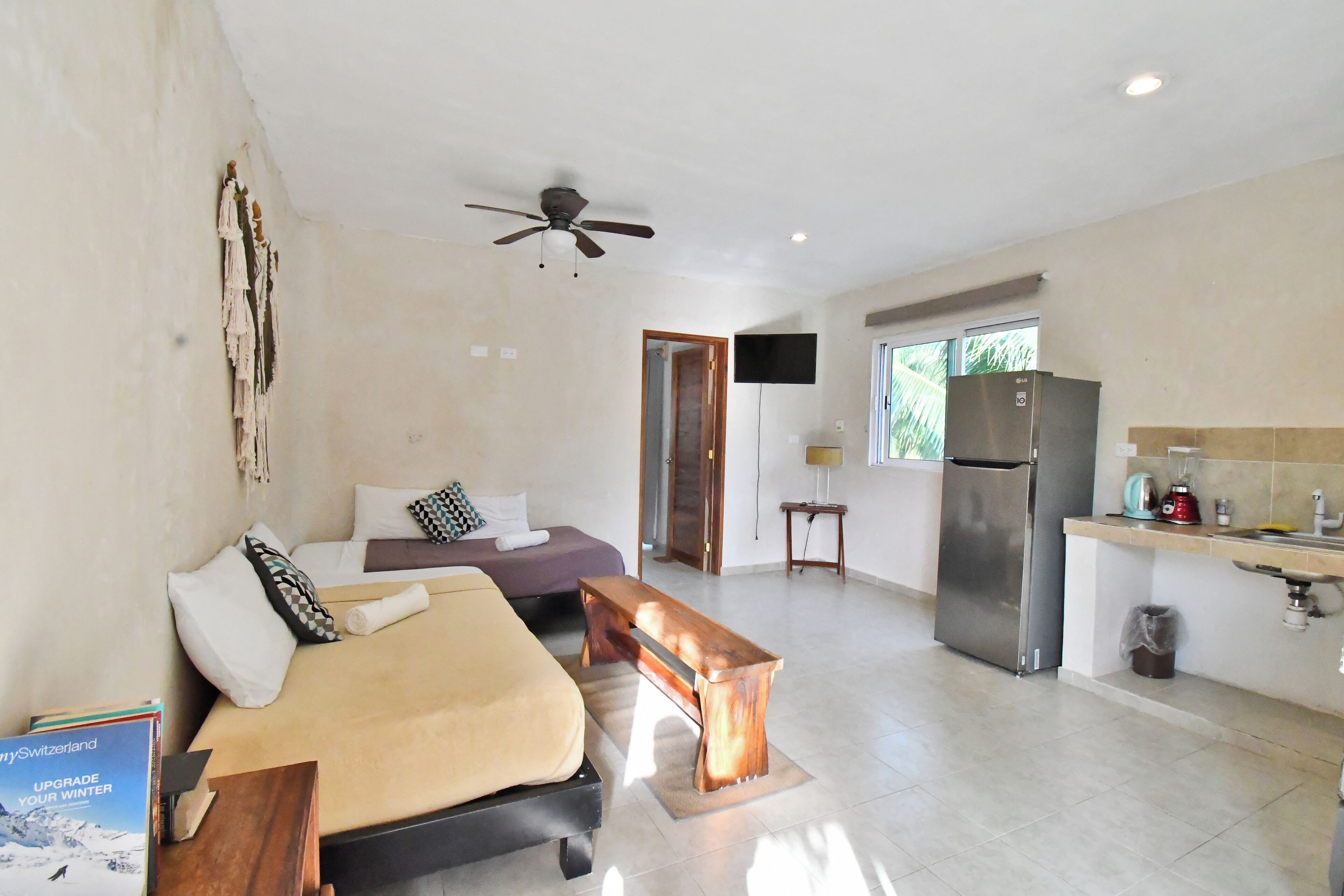 Une chambre d’hôtel moderne équipée d’un lit, d’une banquette, d’un ventilateur de plafond, d’une kitchenette et d’une grande fenêtre.