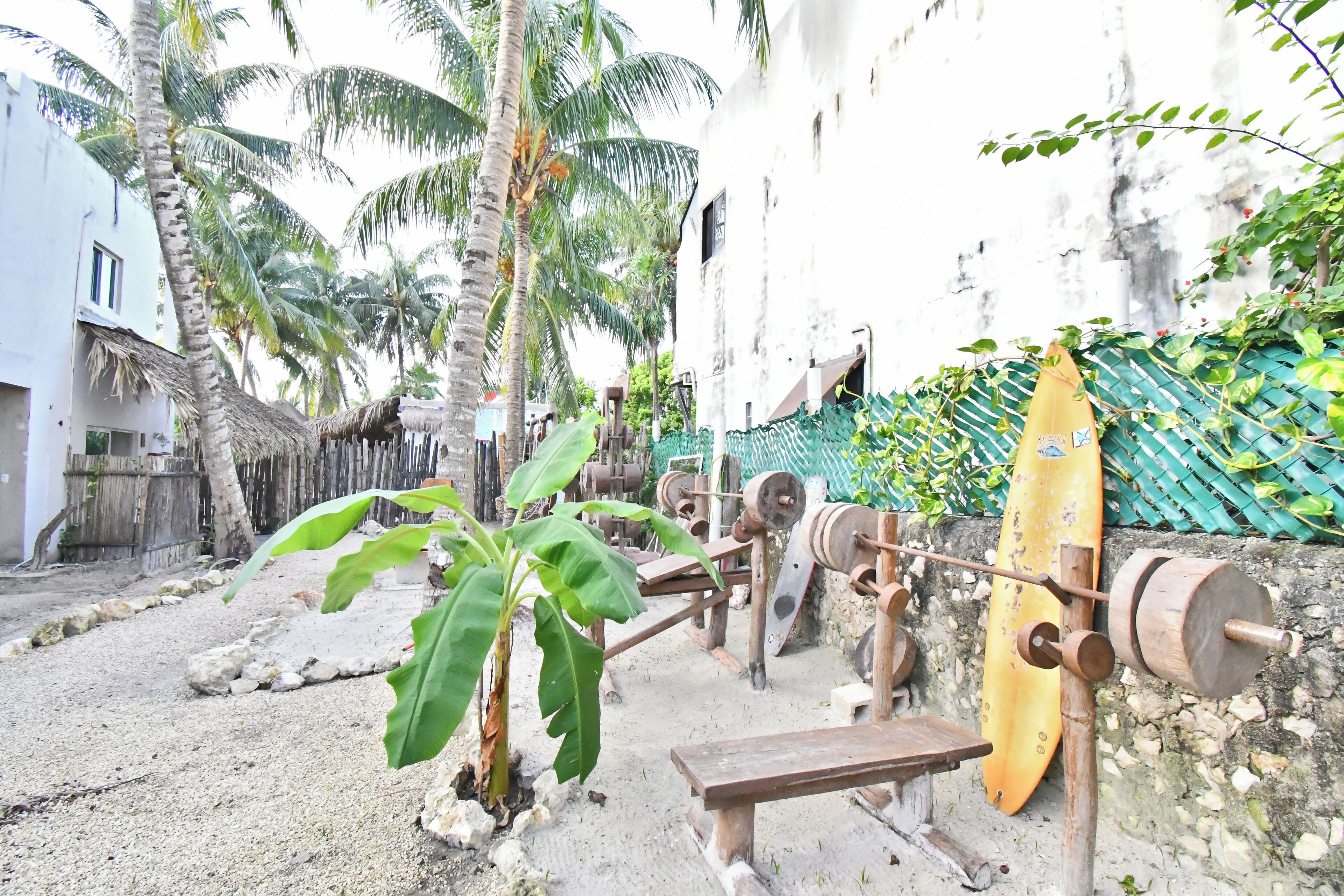 A rustic setting with a traditional water wheel, a wooden bench, and a yellow surfboard leaning against a wall.