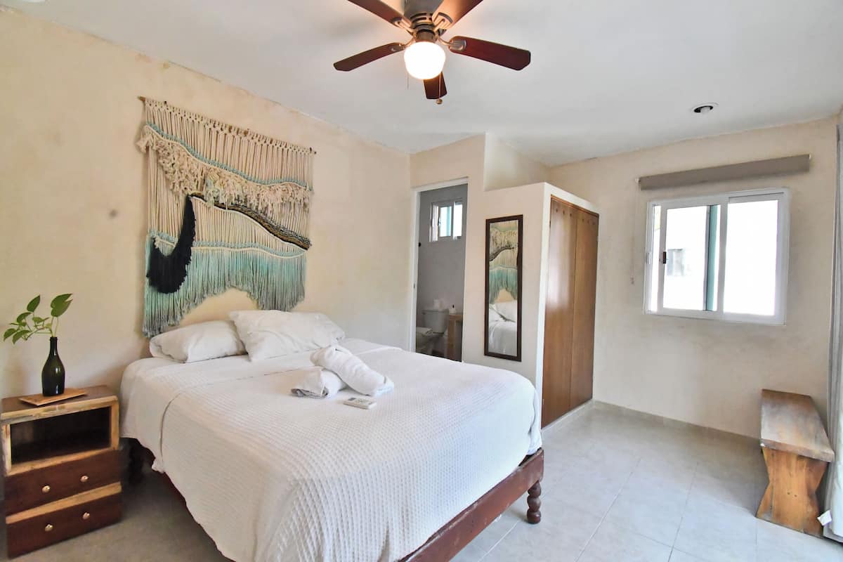 Un dormitorio con cama, ventilador de techo, tapiz en la pared, mesita de noche con planta y ventana.