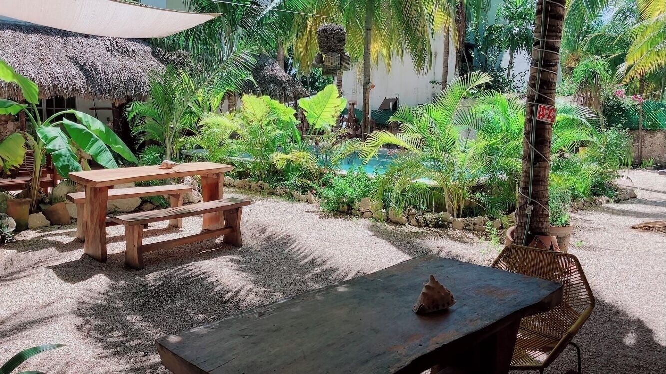 Un'area salotto all'aperto tropicale con tavoli e panche in legno, immersa nel verde e caratterizzata da una struttura con tetto di paglia.