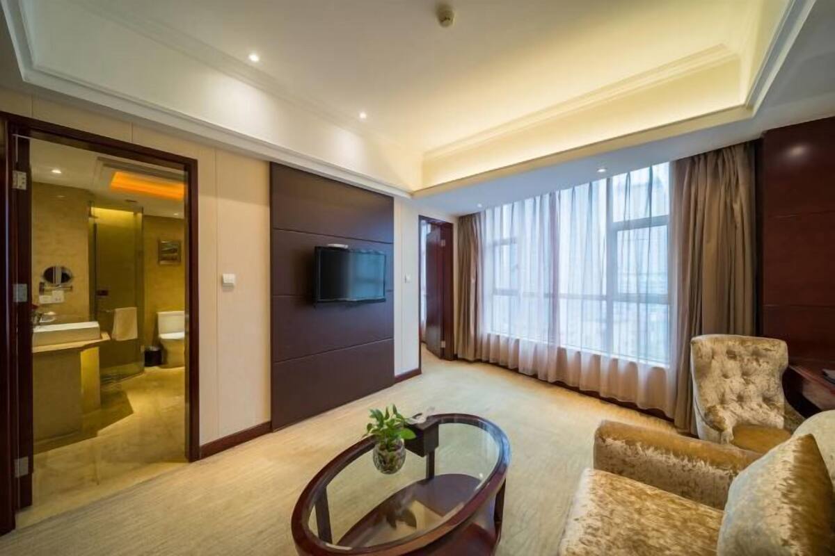 A hotel room with a TV mounted on the wall, a glass coffee table, a plush armchair, and a large window with curtains.
