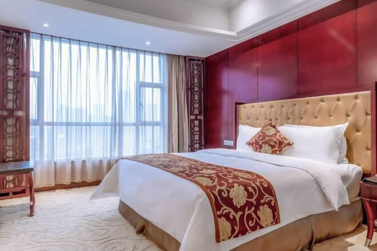 A hotel room with a large bed, a wooden nightstand, and a view of the city through large windows.