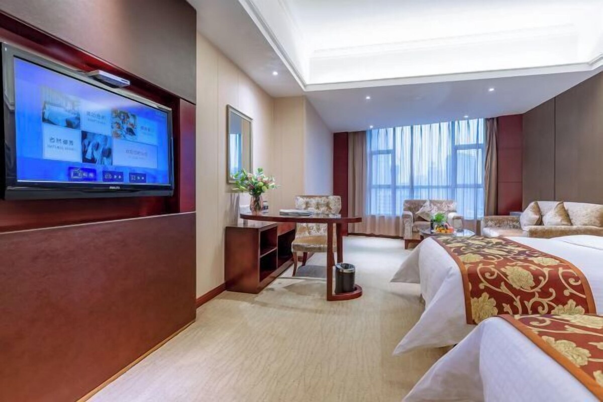 A hotel room with a flat-screen TV mounted on the wall, a bed with a decorative bedspread, a sofa, a small table with a vase of flowers, and a large window with curtains.