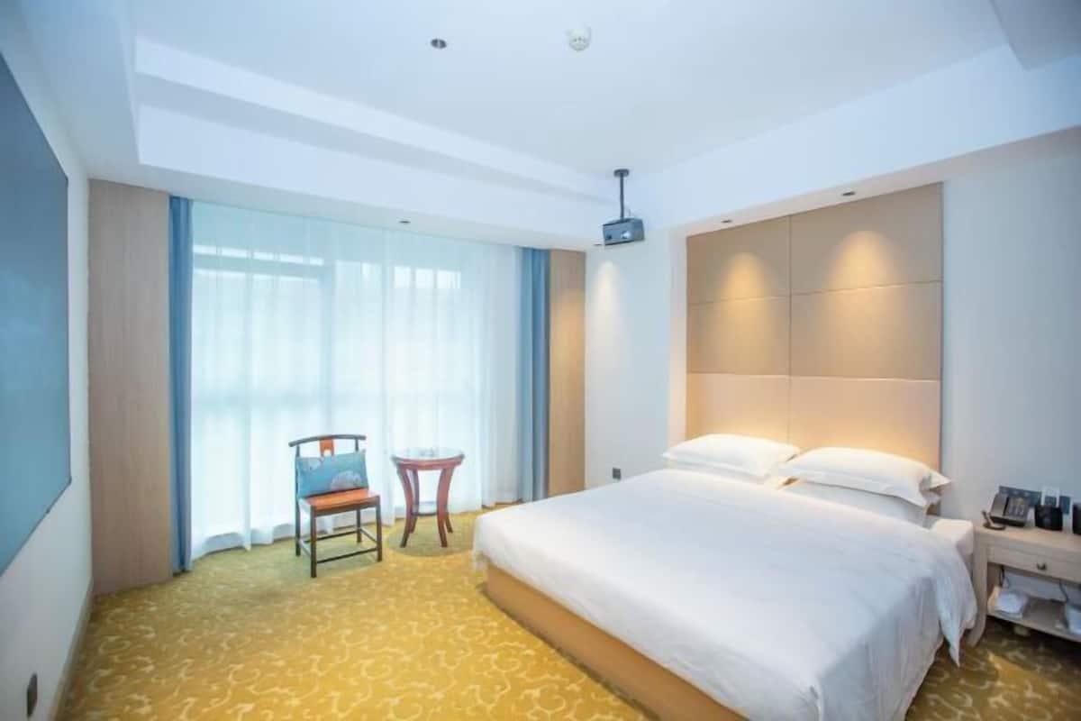 A hotel room with a large bed, a small table, a chair, and a ceiling projector.