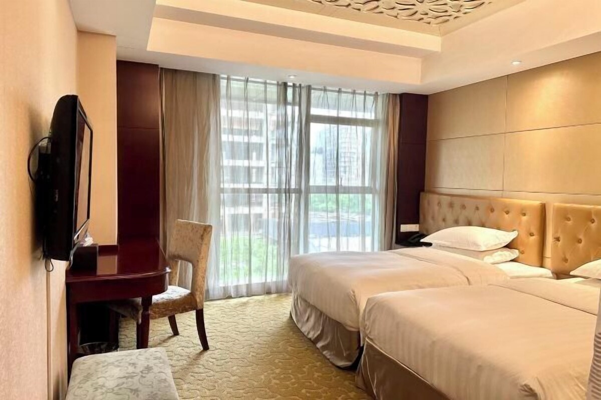 A hotel room with two beds, a desk, a chair, a television, and large windows with curtains.