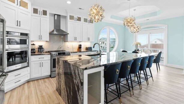 A modern kitchen with white cabinets, a marble island, and a dining area with bar stools. - Beach Access (Ocean Isle Beach)