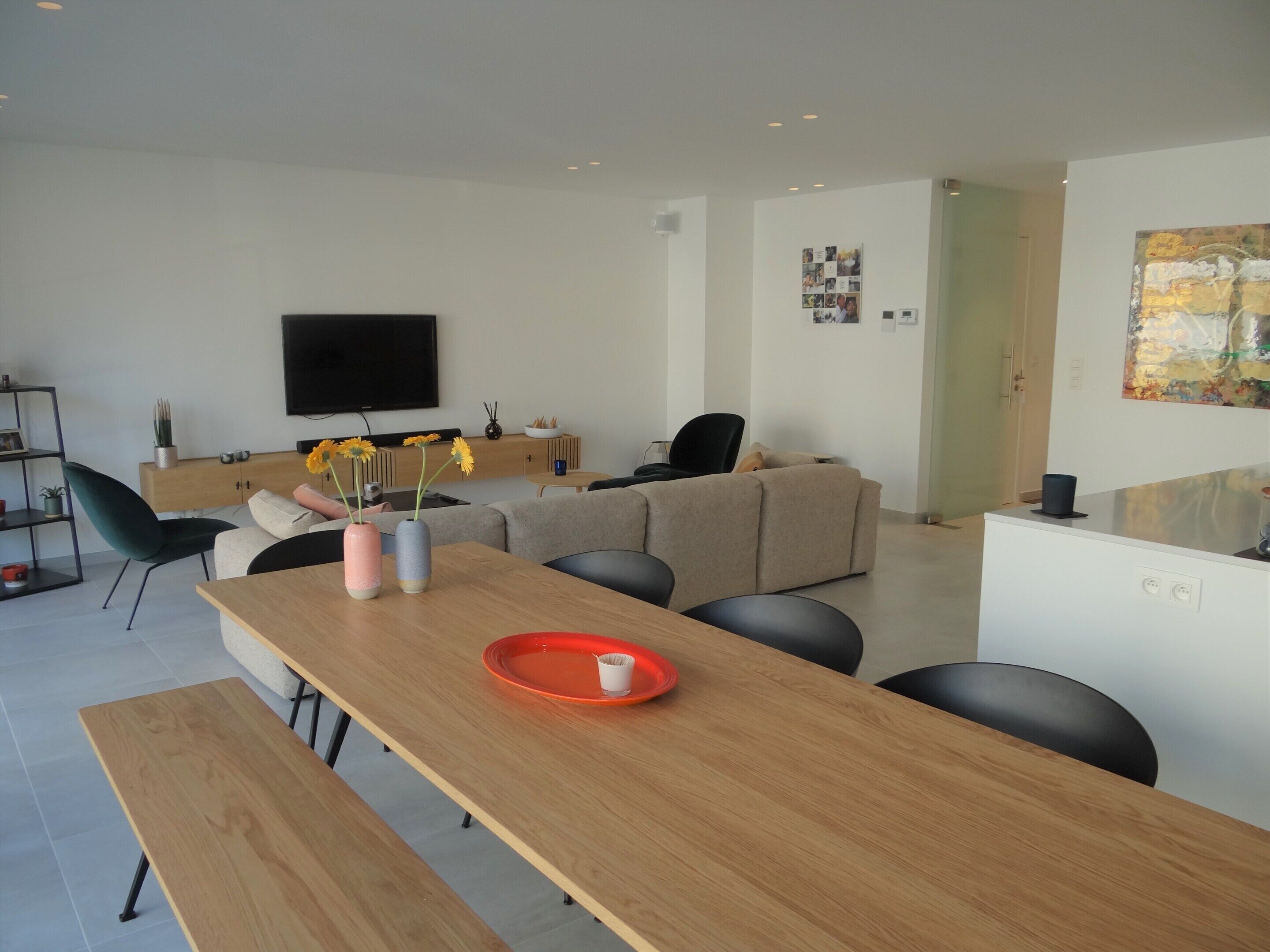 Uma sala de estar moderna com uma mesa de jantar de madeira, um sofá e uma televisão na parede.