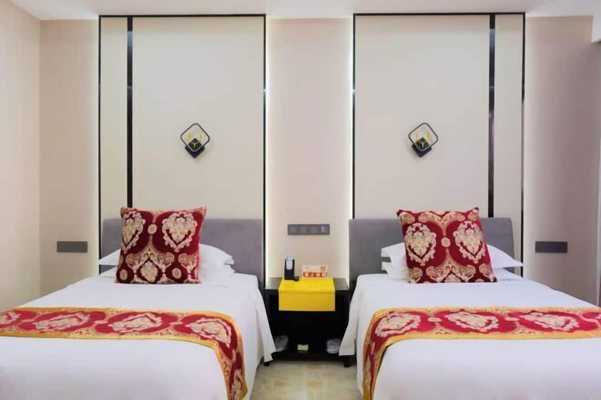 Habitación de hotel con dos camas, cada una con una colcha estampada y almohadas con forma de corazón. Hay una mesita de noche entre las camas con una bandeja amarilla y una lámpara de mesa pequeña.