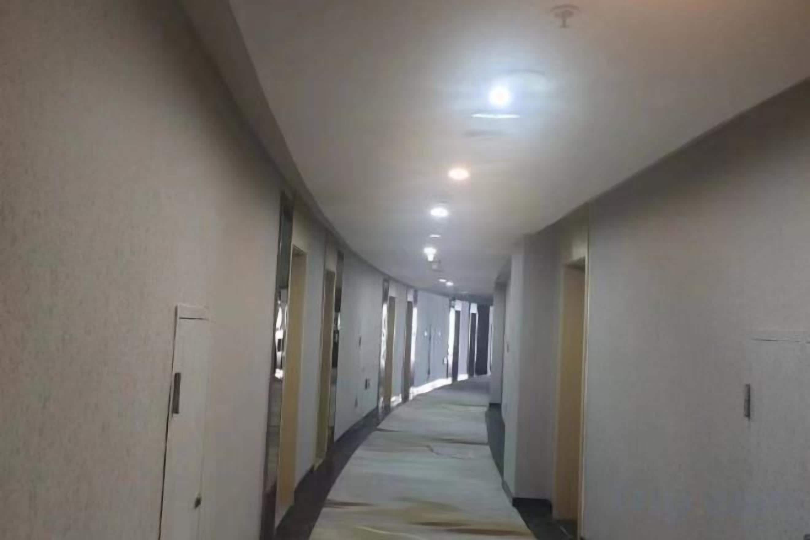 A long, narrow hotel corridor with doors on both sides, recessed lighting, and a carpeted floor.