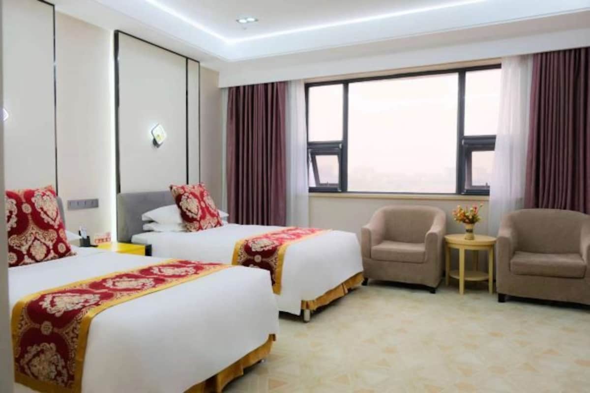 Una habitación de hotel con dos camas, un área de estar con dos sillones, una mesita con un florero y un ventanal con cortinas.