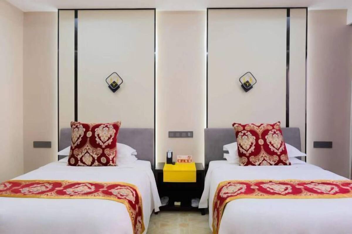 Habitación de hotel con dos camas, cada una con una colcha estampada y almohadas a juego. Entre las camas hay una mesita con un mantel amarillo, y una mesita de noche con una lámpara y una caja de pañuelos.