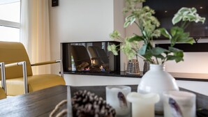 Moderni olohuone, jossa on takka, keltainen nojatuoli, kynttilöillä ja maljakolla koristeltu sohvapöytä sekä seinälle asennettu taulutelevisio.