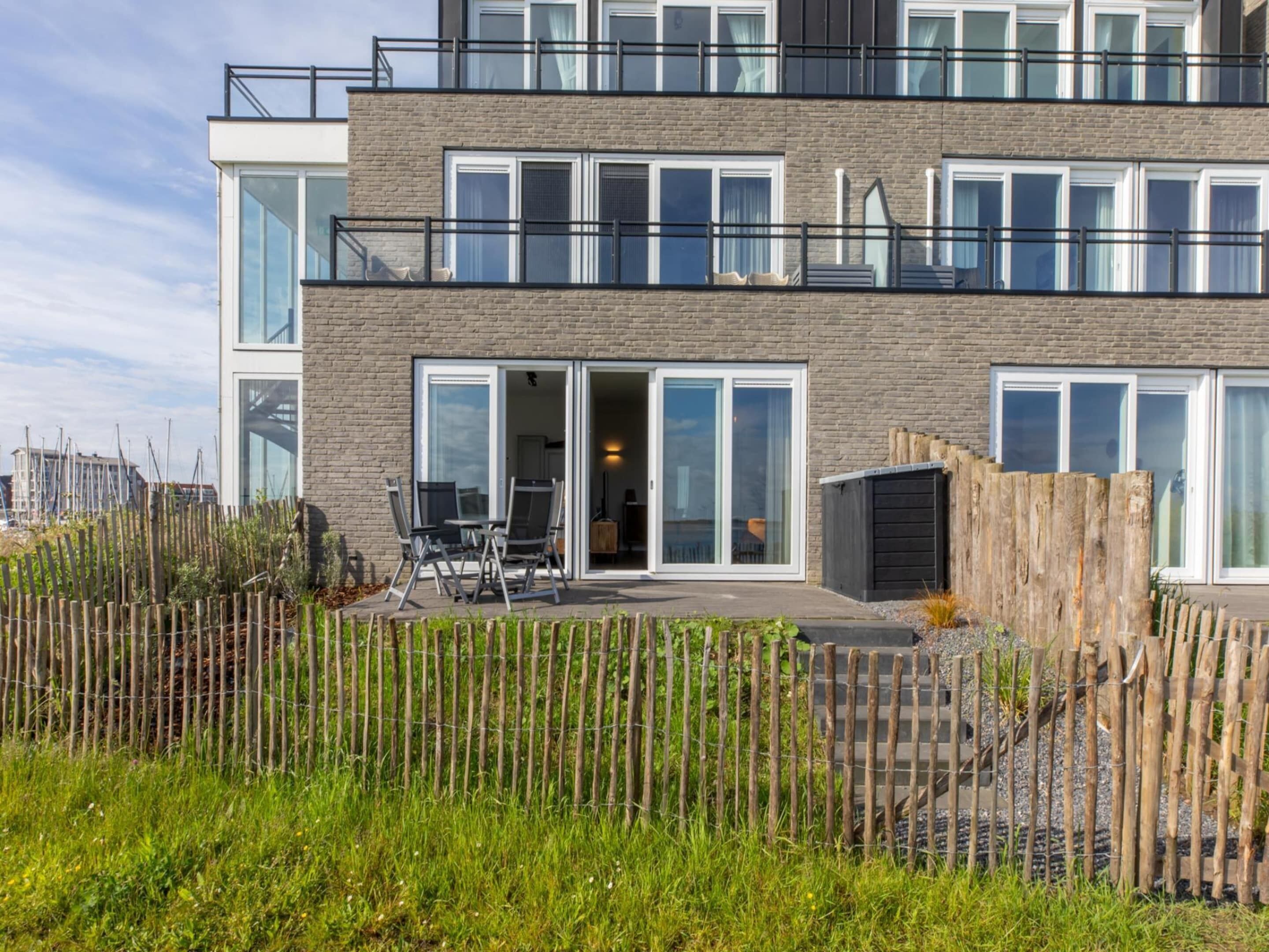 A modern two-story building with large windows, a balcony, and a fenced outdoor area with chairs.