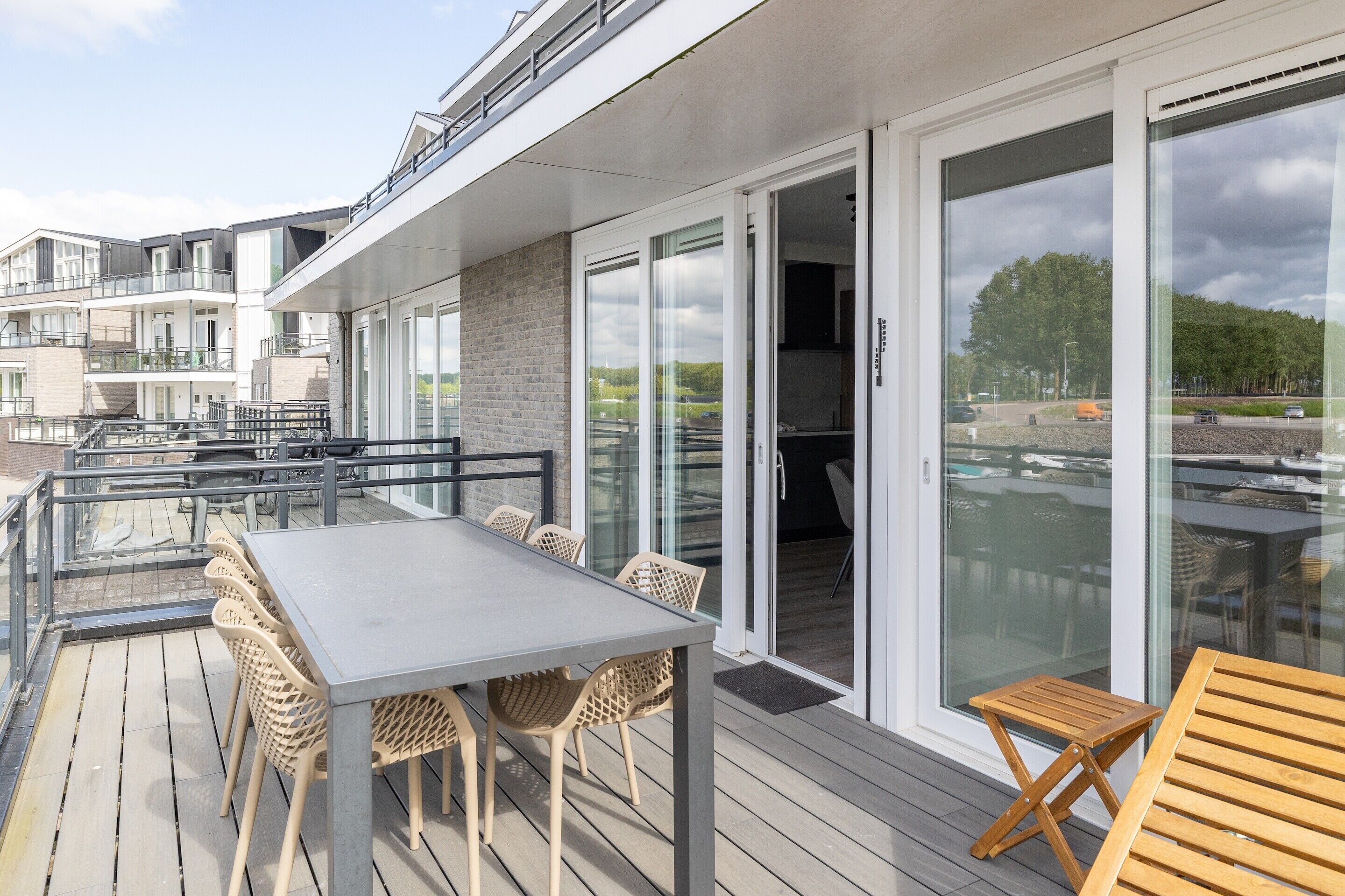 A modern outdoor deck with a glass balustrade, a dining table with chairs, and a wooden lounge chair.