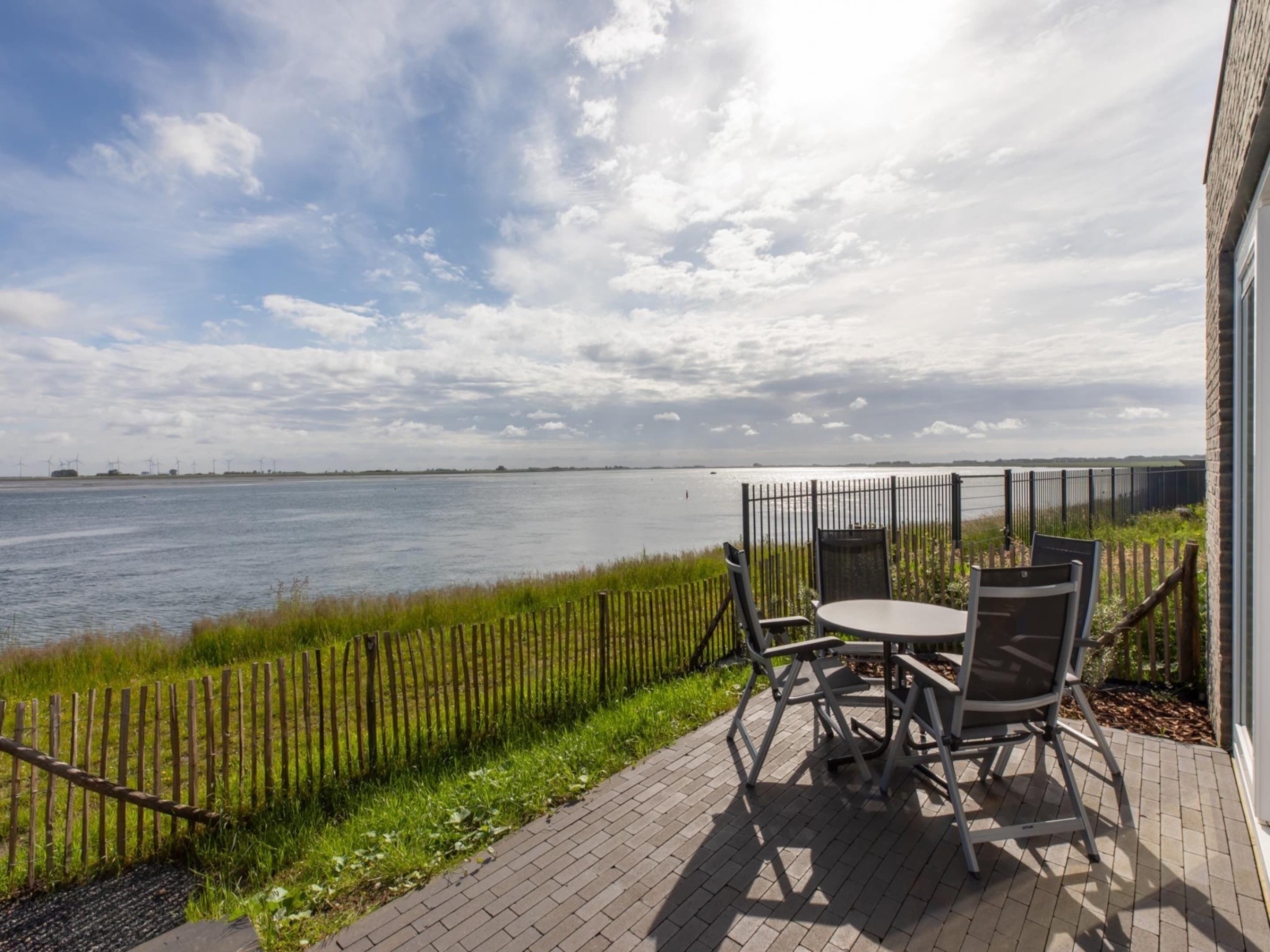 Een terras buiten met een tafel en stoelen, met uitzicht op een wateroppervlak, een houten omheining en een gazon.