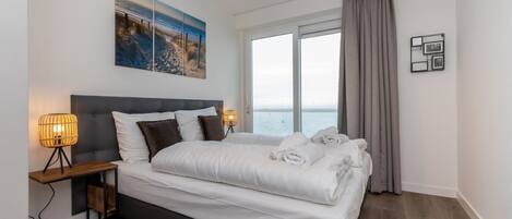 A bedroom with a large bed, a nightstand with a lamp, a view of the sea through a window, and a painting on the wall.