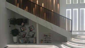 Uma escadaria moderna com corrimão de vidro, rodeada por estantes de livros e plantas em vasos.