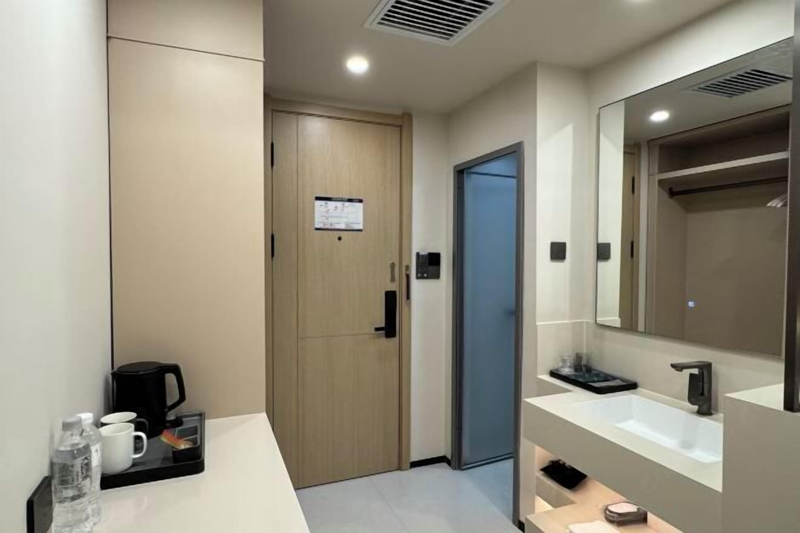 A modern hotel room with a wooden door, a blue elevator, a large mirror, a white sink, and a coffee maker on a tray.