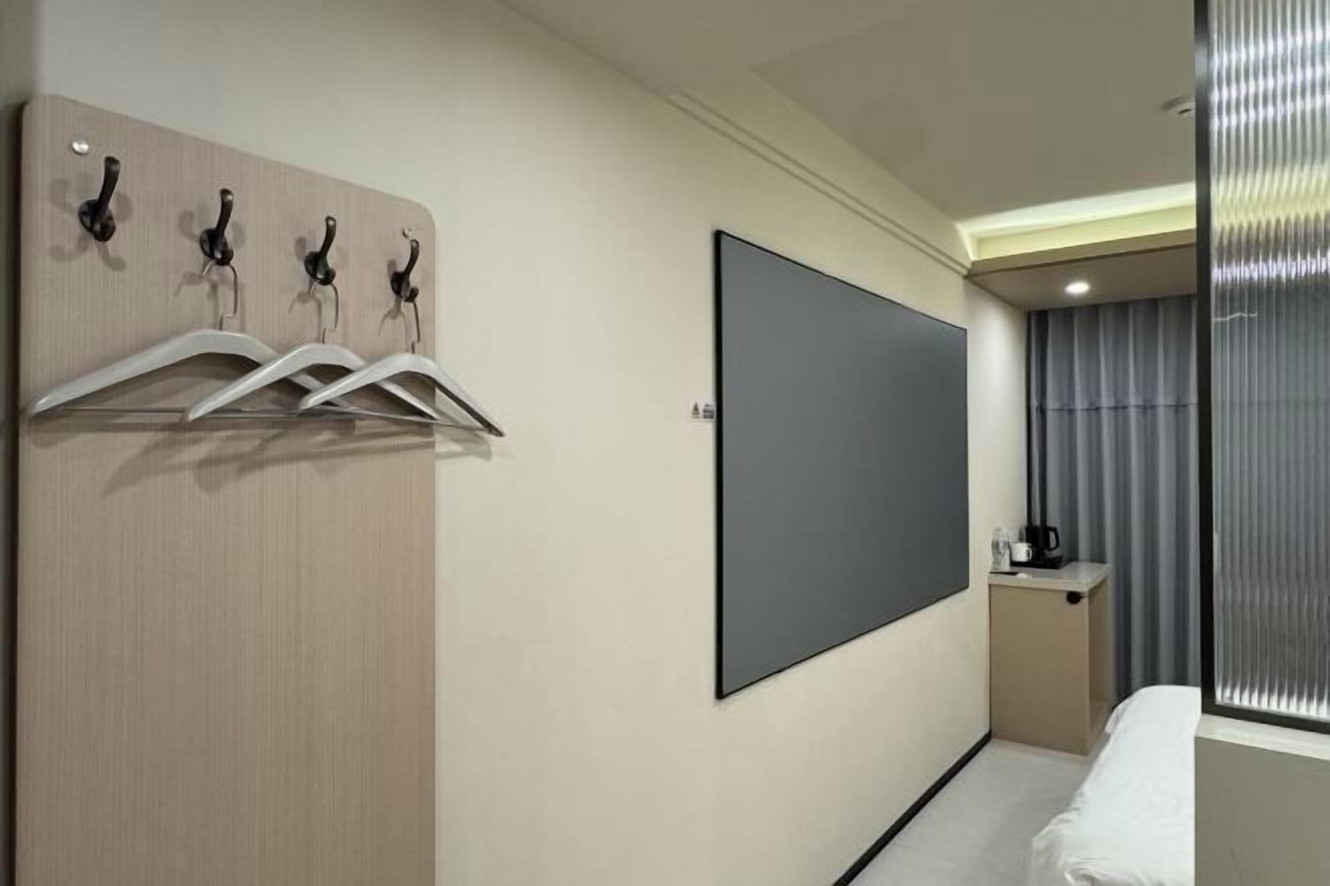Habitación de hotel con cama, televisión montada en la pared, perchero y una pequeña mesa de trabajo con cafetera.