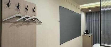 Habitación de hotel con cama, televisión montada en la pared, perchero y una pequeña mesa de trabajo con cafetera.