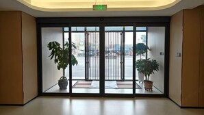 Um saguão de hotel com entrada por porta de vidro, duas plantas em vasos e vista para a rua.