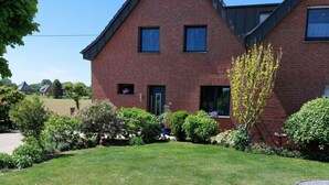 A two-story brick house with a sloped roof, a small front porch, and a well-maintained garden with various shrubs and a tree. - Garden Magic Comfortable Holiday Residence (Goch)