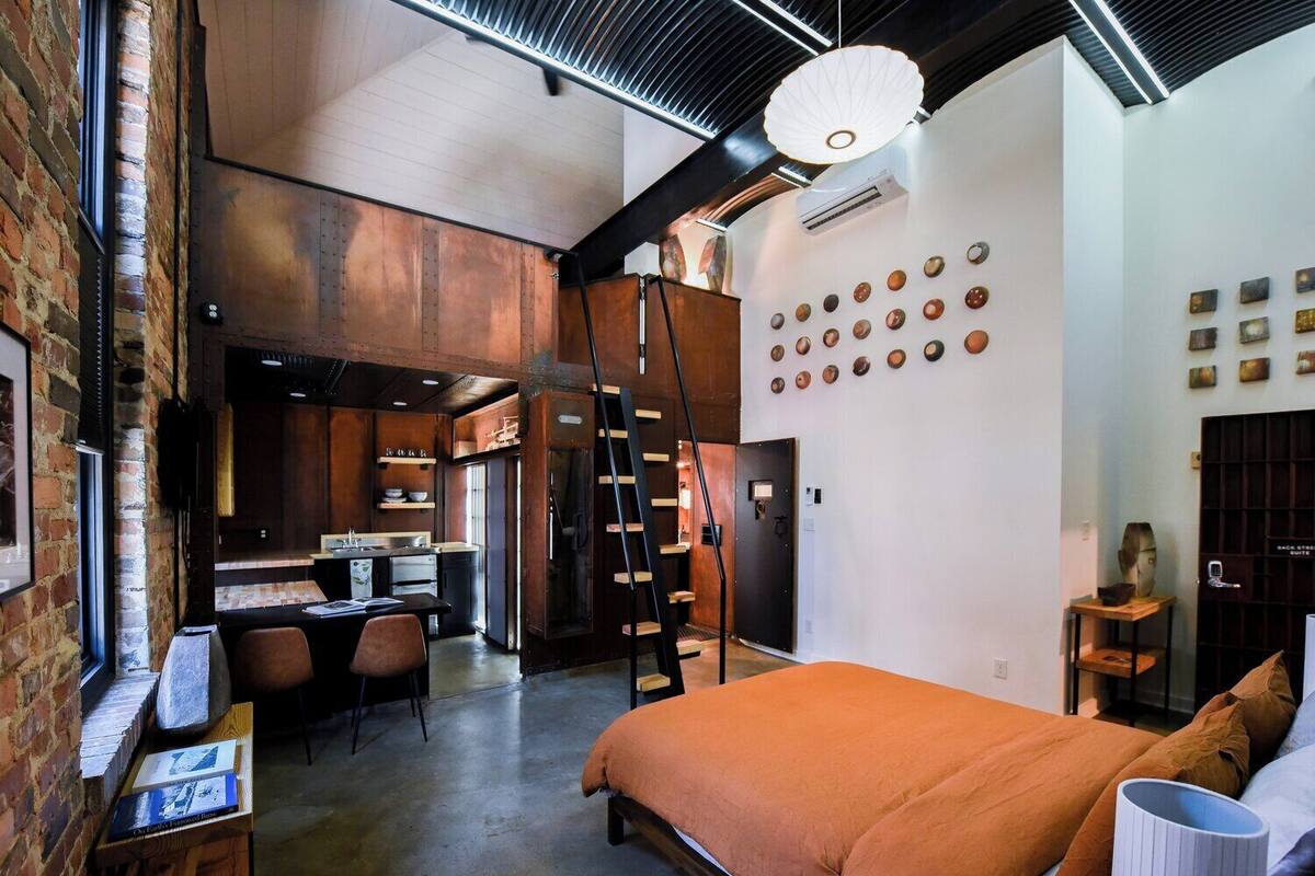 A modern bedroom with a large bed, a desk, and a chair. There is a kitchen area with a stove and a refrigerator.