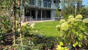 Una casa moderna de dos pisos con un balcón, rodeada por un jardín cuidado y árboles.