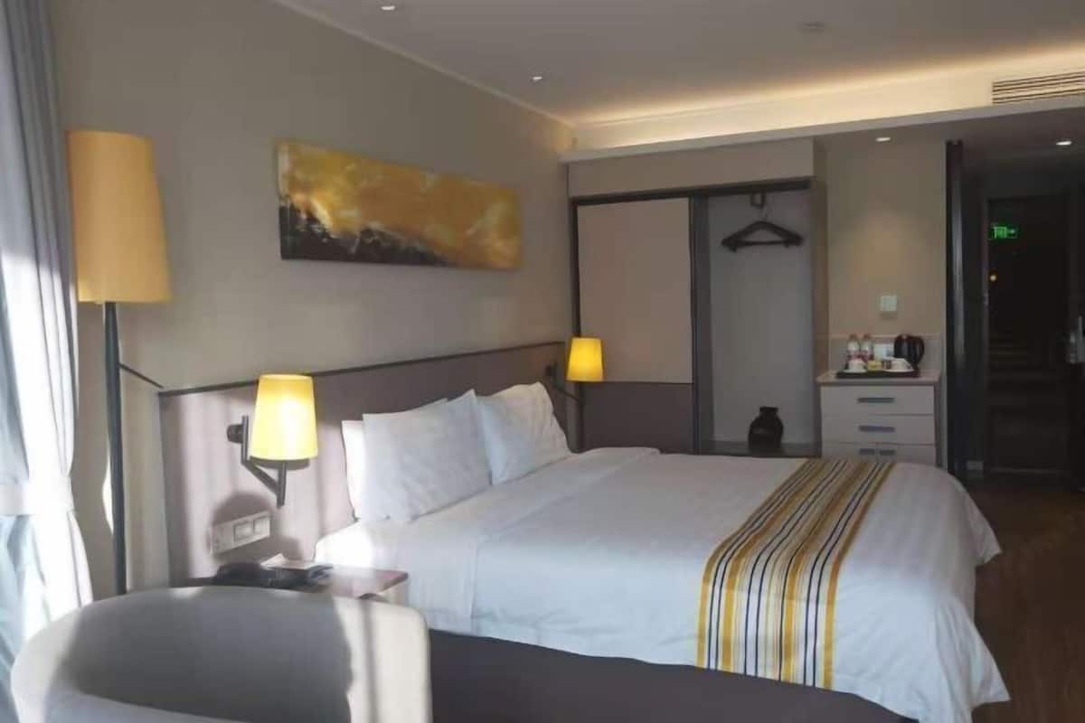 A hotel room with a bed, bedside lamps, a painting, a wardrobe, and a desk with a lamp.
