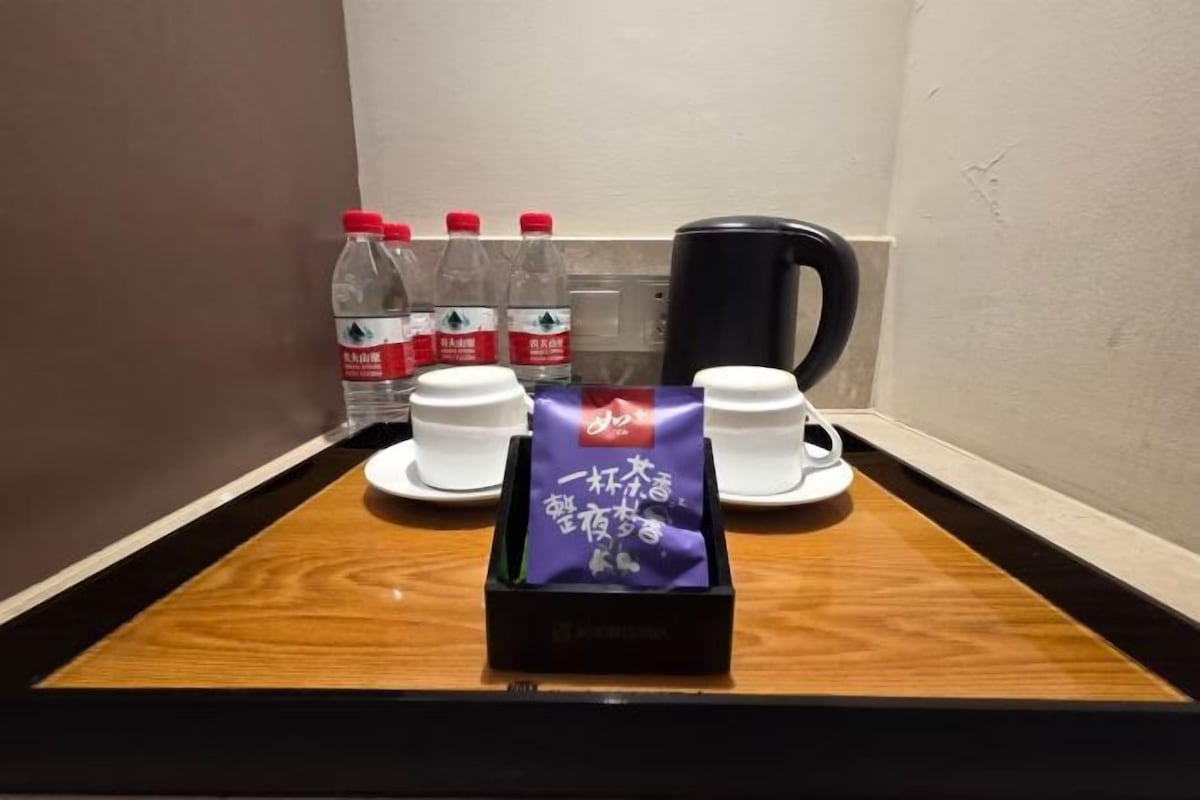 A hotel room with a wooden tray table, two white cups, a black coffee maker, and four water bottles.