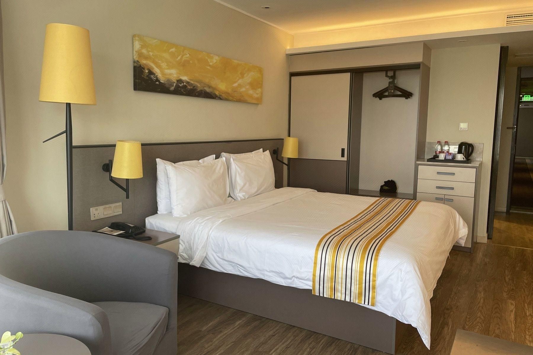 Una habitación de hotel con cama, lámparas de noche, escritorio, silla, perchero y un cuadro en la pared.