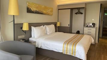 Una habitación de hotel con cama, lámparas de noche, escritorio, silla, perchero y un cuadro en la pared.