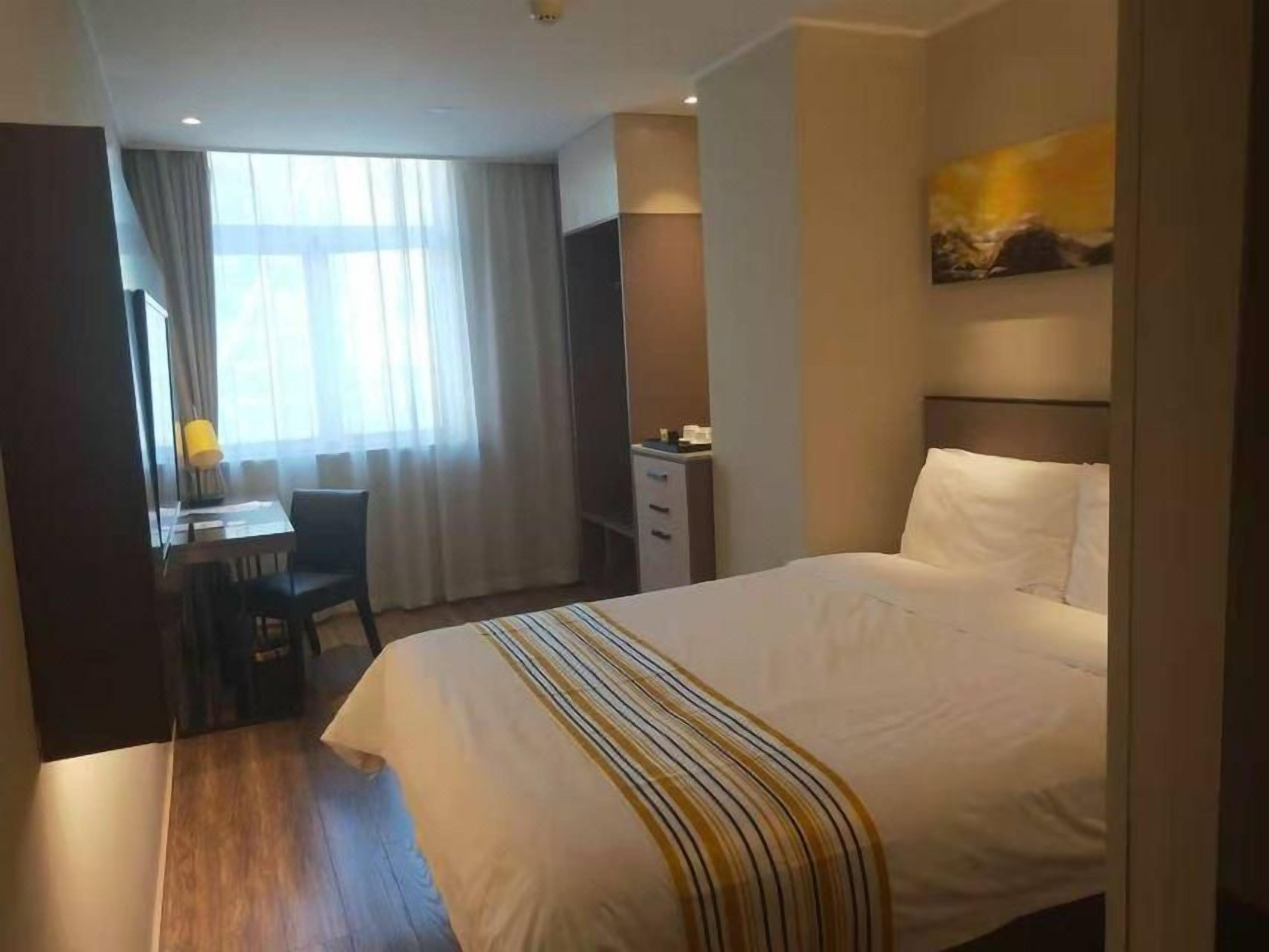 A hotel room with a bed, a desk, a chair, a dresser, and a window with curtains.